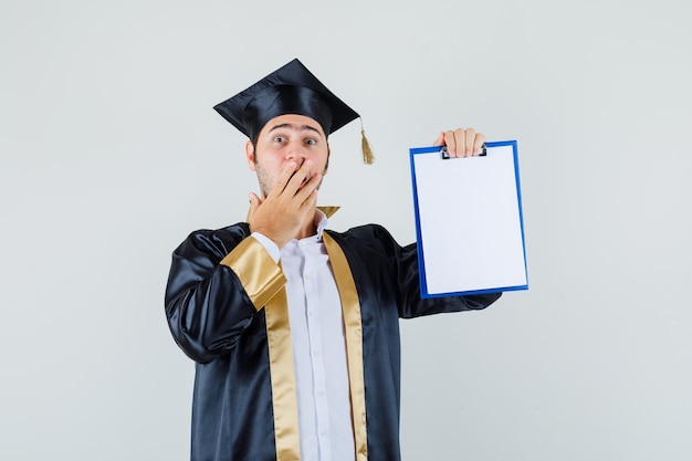 Jeune homme montrant le presse-papiers en uniforme diplômé et à la choqué. vue de face.