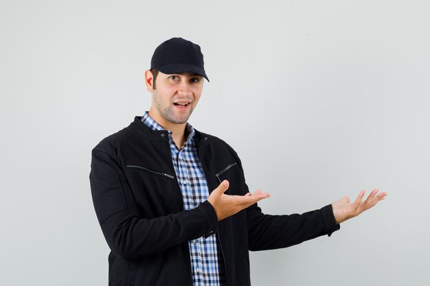 Jeune homme montrant quelque chose ou accueillant en chemise, veste, casquette et à la douce vue de face.