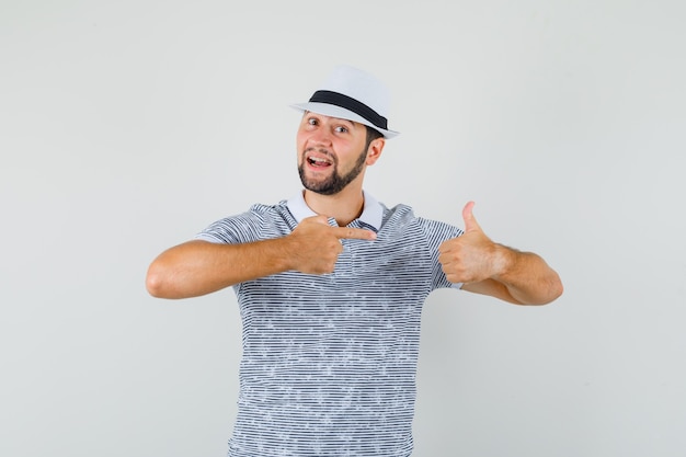 Jeune homme montrant sa main tout en montrant le pouce vers le haut en t-shirt, chapeau et à la vue de face, concentré.