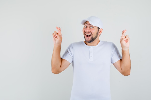 Jeune homme montrant ses doigts croisés en t-shirt blanc, casquette et à la joyeuse. vue de face.