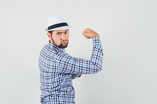 Jeune homme montrant ses muscles du bras en chemise à carreaux, chapeau et à la recherche résolue.