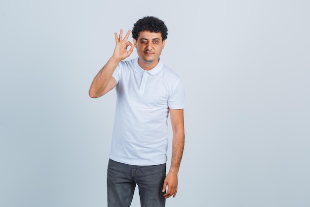 Jeune homme montrant un signe ok et un clin d'œil en t-shirt blanc et jeans et l'air heureux. vue de face.