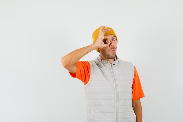 Jeune homme montrant le signe ok sur les yeux en t-shirt, veste, chapeau et à la curiosité.