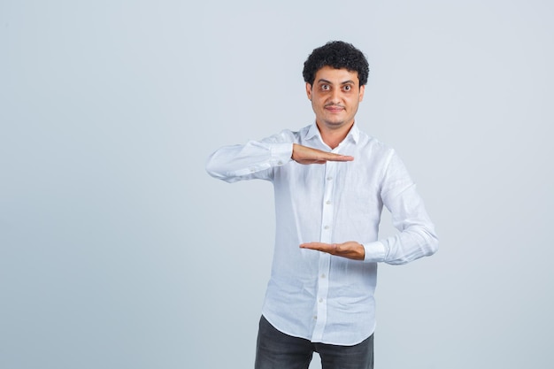 Jeune homme montrant le signe de la taille en chemise blanche, pantalon et à l'air confiant, vue de face.