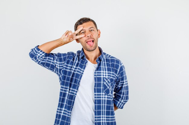 Jeune homme montrant le signe de la victoire près des yeux en chemise et à la recherche amusée. vue de face.