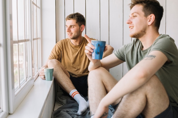 Jeune homme montrant son ami par la fenêtre tout en tenant une tasse de café