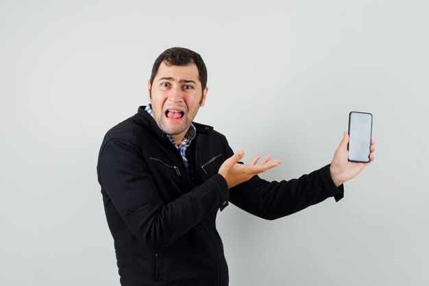 Jeune homme montrant un téléphone mobile en chemise, veste et à la perplexité, vue de face.