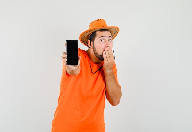 Jeune homme montrant un téléphone mobile en t-shirt orange, chapeau et à la surprise, vue de face.