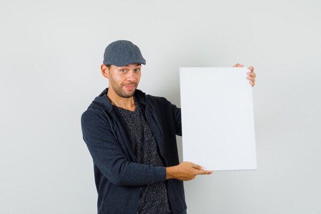 Jeune homme montrant une toile vierge et souriant en t-shirt, veste, vue de face de casquette.