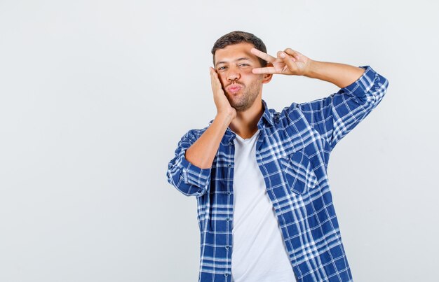 Jeune homme montrant v-sign près des yeux avec la main sur la joue en chemise et à la vue de face, joyeuse.