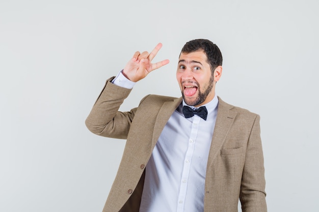 Jeune homme montrant v-sign, qui sort la langue en costume et à la recherche d'énergie. vue de face.