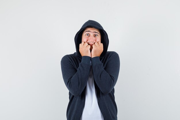 Jeune homme mordant les poings émotionnellement en t-shirt, veste et à la peur. vue de face.