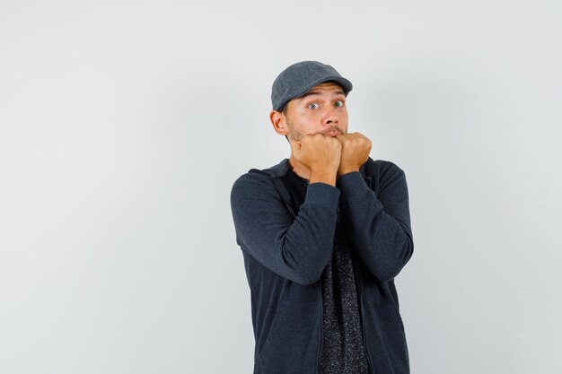 Jeune homme mordre les poings émotionnellement en t-shirt, veste, casquette et à la peur, vue de face.