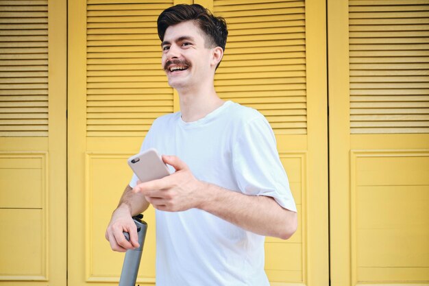 Jeune homme moustache attrayant en T-shirt blanc tenant joyeusement un téléphone portable en se tenant debout avec un fond jaune sur la rue de la ville