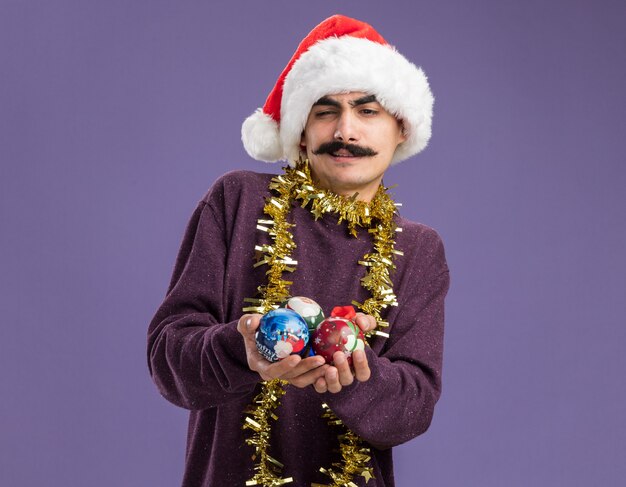 Jeune homme moustachu portant un bonnet de noel de noël avec des guirlandes autour du cou tenant des boules de noël avec un sourire sceptique debout sur un mur violet