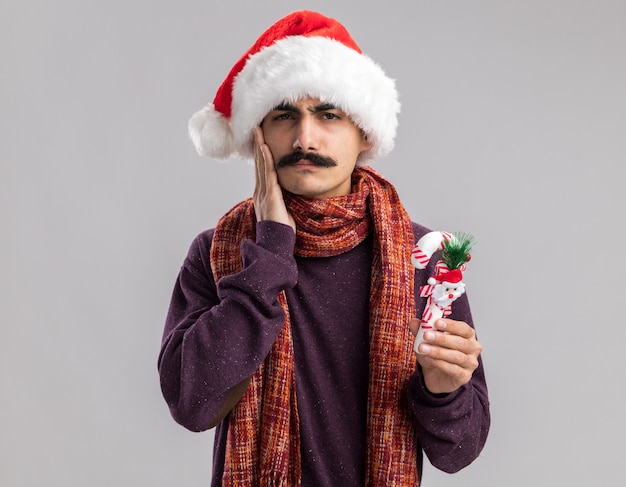 Jeune homme moustachu portant chapeau de Père Noël avec foulard chaud autour de son cou tenant la canne à sucre de Noël à la confusion