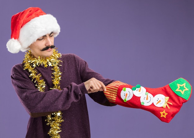 Photo gratuite jeune homme moustachu portant chapeau de père noël avec guirlandes autour du cou tenant des bas de noël à la confusion debout sur fond violet