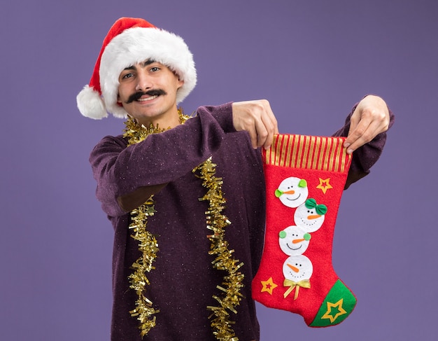 Photo gratuite jeune homme moustachu portant chapeau de père noël avec guirlandes autour du cou tenant des bas de noël regardant la caméra avec un visage heureux souriant joyeusement debout sur fond violet