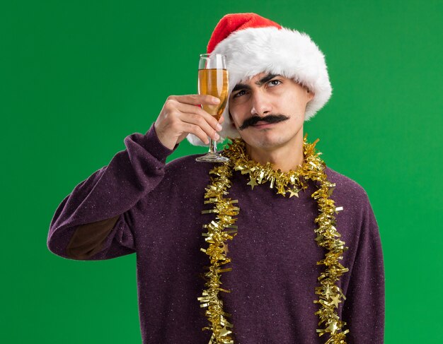 Jeune homme moustachu portant chapeau de Père Noël avec des guirlandes autour du cou tenant un verre de champagne à la perplexité