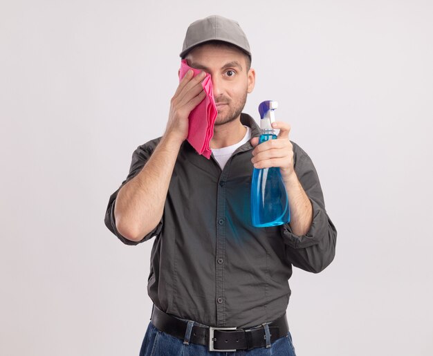 Jeune homme de nettoyage portant des vêtements décontractés et cap tenant le spray de nettoyage et un chiffon en essuyant ses yeux avec un chiffon debout sur un mur blanc