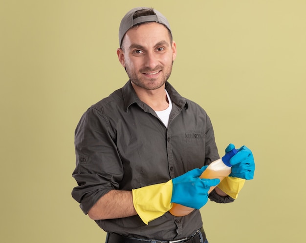 Jeune homme de nettoyage portant des vêtements décontractés et capuchon dans des gants en caoutchouc tenant une bouteille avec des produits de nettoyage à la recherche de sourire sur le visage debout sur un mur vert
