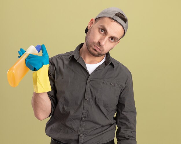 Photo gratuite jeune homme de nettoyage portant des vêtements décontractés et capuchon en gants en caoutchouc tenant une bouteille avec des produits de nettoyage à la fatigue et dérangé debout sur le mur vert