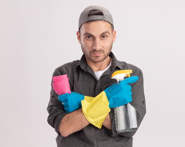 Jeune homme de nettoyage portant des vêtements décontractés et une casquette de gants en caoutchouc tenant un spray de nettoyage et un chiffon à la recherche de visage sérieux debout sur un mur orange