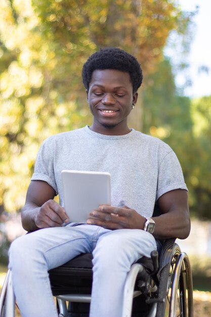 Jeune homme noir en fauteuil roulant passant du temps à l'extérieur dans le parc, lisant une tablette numérique, souriant et s'amusant le jour d'automne ensoleillé. De plein fouet. Technologies modernes, temps libre, concept de handicap.