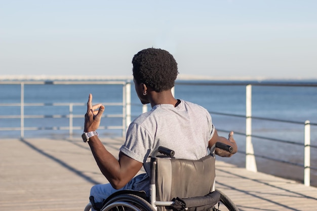 Photo gratuite jeune homme noir en fauteuil roulant s'amusant au bord de la mer, rappant et bougeant ses mains. afro-américain handicapé physique s'amusant à l'extérieur. jeunesse, activité de loisirs, concept de handicap.