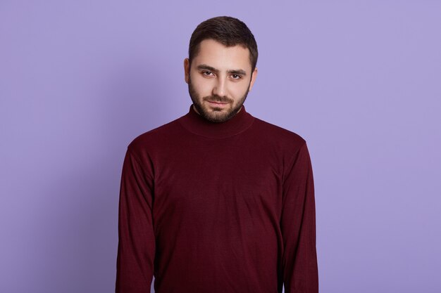 Jeune homme non rasé portant un pull bordeaux posant sur fond violet avec une expression faciale grave