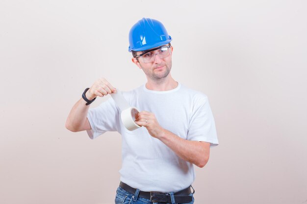 Jeune homme ouvrant un rouleau de ruban adhésif en t-shirt, jeans, casque et regardant attentivement