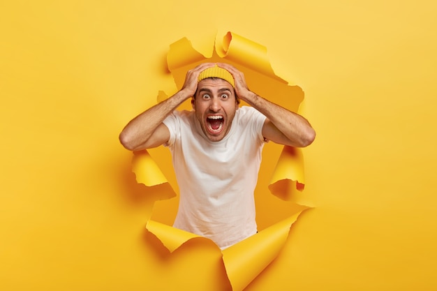 Jeune homme paniqué émotionnel garde les deux mains sur la tête, crie fort, regarde avec des yeux buggés, porte un t-shirt blanc