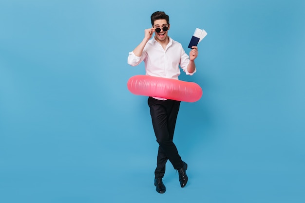 Jeune homme en pantalon noir et chemise blanche pose joyeusement avec anneau en caoutchouc. Portrait en pied de gars avec passeport et billets pour le repos.