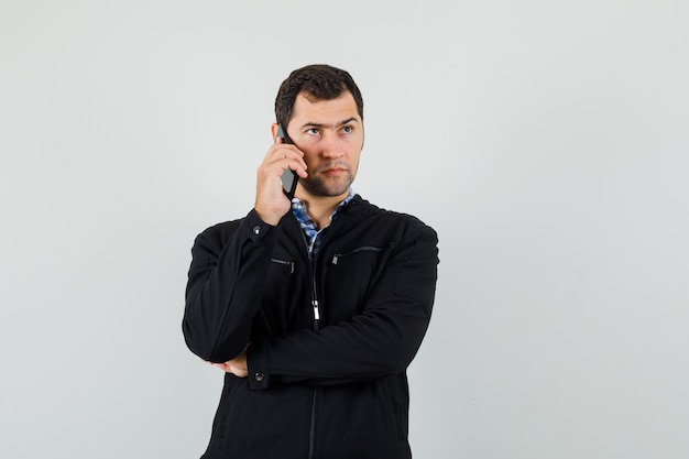 Jeune homme parlant au téléphone mobile en chemise, veste et regardant pensif, vue de face.