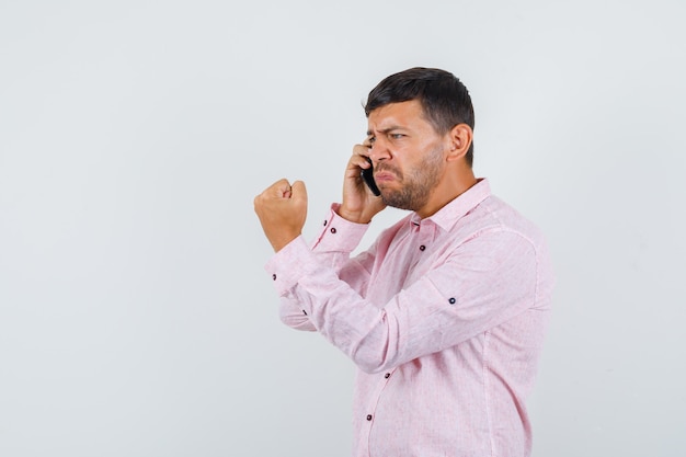 Jeune homme parlant au téléphone mobile avec le poing fermé en chemise rose et à la vue de face, nerveux.