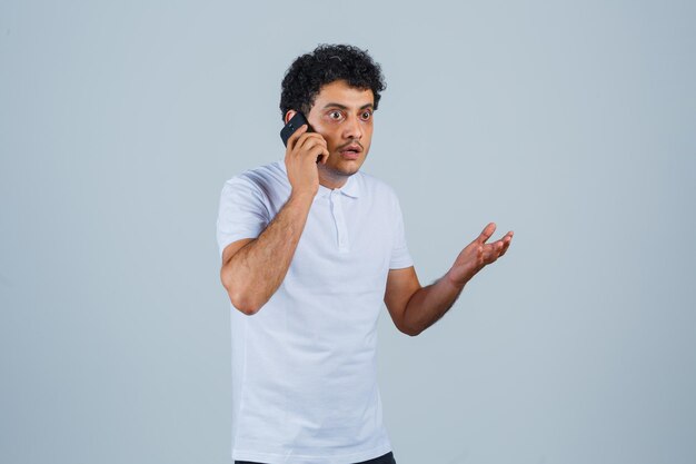 Jeune homme parlant au téléphone mobile en t-shirt blanc et à la perplexité, vue de face.