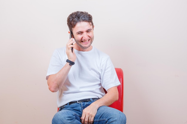 Jeune homme parlant au téléphone portable alors qu'il était assis sur une chaise en t-shirt, jeans et l'air heureux