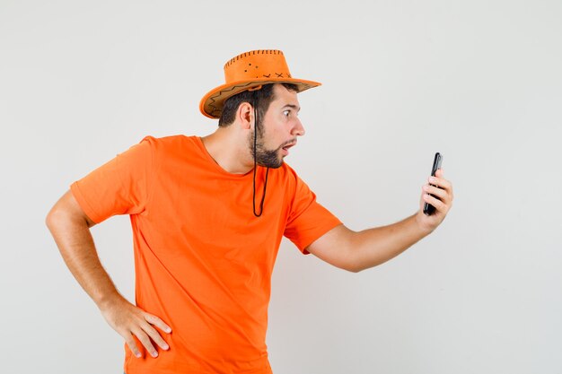 Jeune homme parlant sur le chat vidéo en t-shirt orange, chapeau et à la vue choquée, de face.