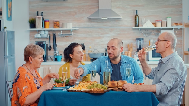 Photo gratuite jeune homme parlant pendant le dîner plusieurs générations, quatre personnes, deux couples heureux discutant et mangeant pendant un repas gastronomique, profitant du temps à la maison, dans la cuisine assis près de la table.