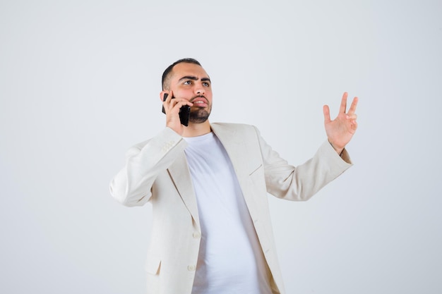 Jeune homme parlant à quelqu'un par téléphone en t-shirt blanc, veste et semblant sérieux, vue de face.