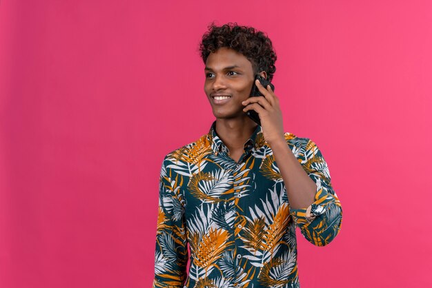 Un jeune homme à la peau foncée beau avec des cheveux bouclés en chemise imprimée de feuilles de parler avec un téléphone mobile