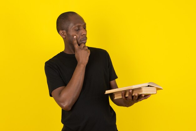 Jeune homme à la peau foncée tenant un livre et pensant sur un mur jaune
