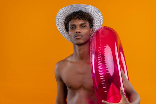 Un jeune homme à la peau sombre et confiant avec des cheveux bouclés portant un chapeau de soleil tenant un anneau de piscine gonflable