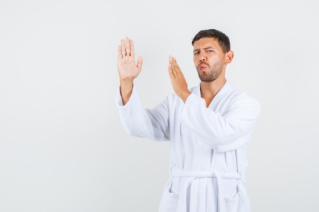 Jeune homme en peignoir blanc montrant le geste de côtelette de karaté, vue de face.