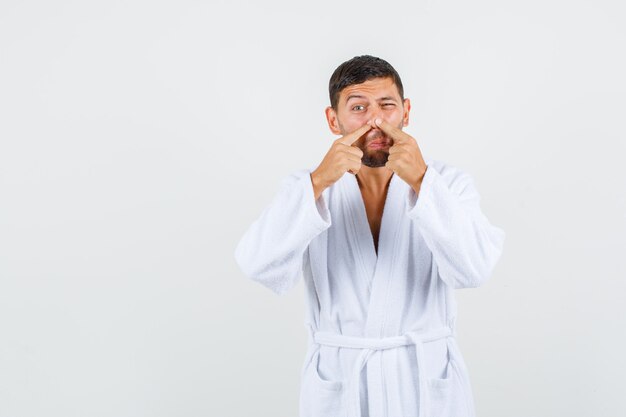 Jeune homme en peignoir blanc serrant l'acné sur son nez, vue de face.