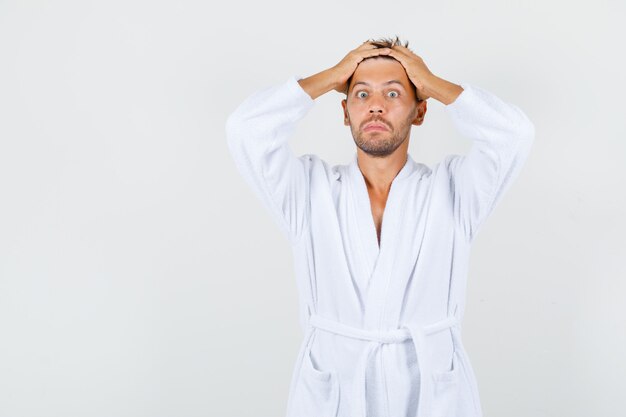 Photo gratuite jeune homme en peignoir blanc tenant les mains sur la tête et à la vue de face, surpris.