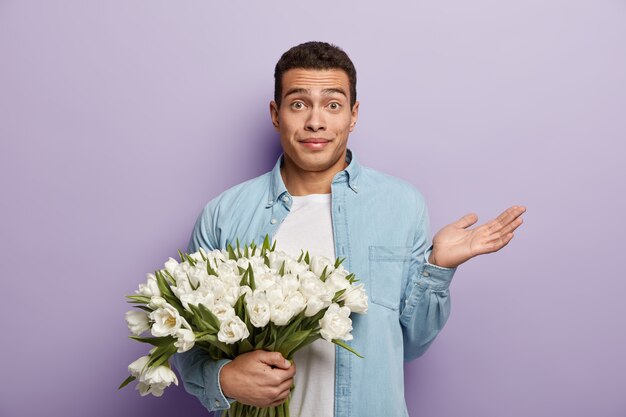 Un jeune homme perplexe tient un gros bouquet de fleurs, lève la main avec indignation, arrive à la date