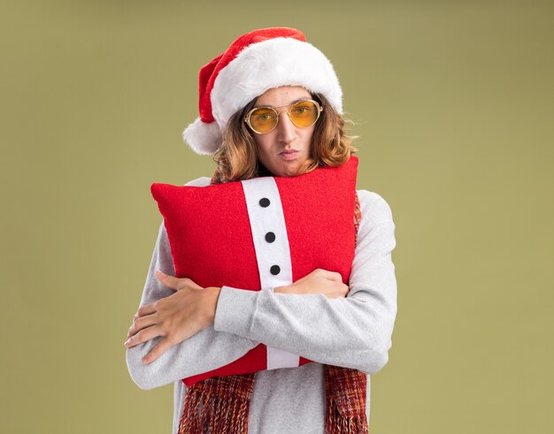 Jeune homme plein de ressentiment portant bonnet de noel de Noël et des lunettes jaunes avec écharpe chaude autour de son cou tenant un oreiller de Noël regardant la caméra pincer les lèvres debout sur fond vert