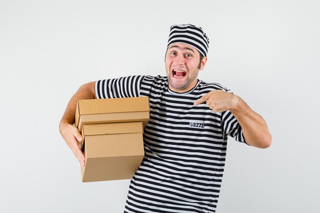 Jeune homme pointant sur des boîtes en carton en t-shirt, chapeau et air heureux. vue de face.