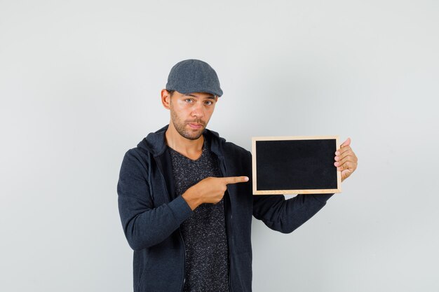 Jeune homme pointant sur le cadre en t-shirt, veste, casquette et à la confiance. vue de face.